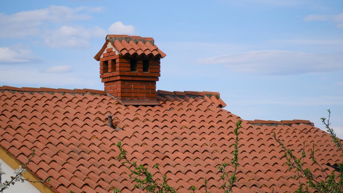 A charming red brick chimney stands atop a detailed clay-tiled roof under a clear blue sky.